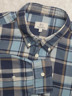 Woolrich rutig skjorta blå storlek L - Rutig skjorta från Woolrich i blått, ljusblått, beige och vitt. Klassisk modell med button down-krage och bröstficka, med ett stilrent rutmönster som passar bra till jeans eller chinos.  Storlek L  Mycket bra begagnat skick.  Mått • Bröstbredd: ca 57 cm • Plagglängd: ca 70 cm • Axelbredd: ca 45 cm • Ärmlängd: ca 64 cm  Material Materiallapp saknas. Tyget bedöms vara bomull, men exakt materialsammansättning är okänd.  Varan är begagnad och säljs i befintligt skick, se bilder för detaljer.