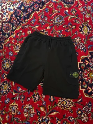 Svarta shorts från Stone Island - Svarta shorts från Stone Island med elastisk midja och snörning. Har en ficka på sidan med den klassiska Stone Island-loggan. Perfekta för sommaren och har en avslappnad passform.