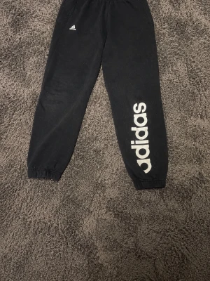 Svarta Adidas mjukisbyxor med tryck - Svarta mjukisbyxor från Adidas med stort vitt logotryck längs ena benet och liten Adidas-logga vid höften. Byxorna har resår i midjan och vid bensluten för en skön passform. Perfekta för chill eller träning.