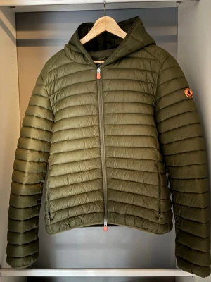 Olivgrön save the duck jacka  - Snygg olivgrön dunjacka med huva och quiltad design. Jackan har dragkedja framtill och en patch på ärmen. Perfekt för kalla vinterdagar och ger en stilren look. Lättviktig och smidig att bära, passar till det mesta.
