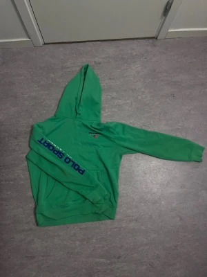 Grön hoodie Polo Sport Ralph Lauren - Snygg grön hoodie från Polo Sport Ralph Lauren med blå logga på ärmen och broderad logga samt amerikansk flagga på bröstet. Klassisk huva och känguruficka framtill. Perfekt för en sportig och avslappnad stil.