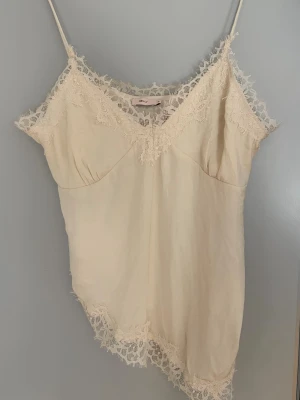 Beige linnetopp med spetsdetaljer - Säljer en beige linnetopp med tunna axelband och vacker spets längs v-ringningen och nederkanten. Toppen har en feminin och romantisk look med asymmetrisk kant och mjukt material. Perfekt för att styla lager på lager eller till jeans.