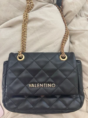 Svart handväska Valentino - Snygg svart  handväska från Valentino med guldfärgade kedjor (men de har rostads en hel del) en av fickorna med dragkedja har vita smuts fläckar, men inget man tänker på. Den är lite sliten, men funkar helt perfekt att använda. Lappen är bortklippt i väskan tyvärr. Köpte på Vinted.