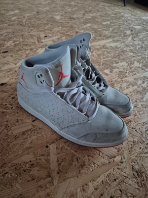 Gråa Air Jordan high sneakers - Säljer ett par gråa Air Jordan sneakers med högt skaft och perforerade sidor. Skorna har röda detaljer som Jumpman-loggan på plösen och sidan. Tillverkade i mockaliknande material med klassisk snörning och vit sula.