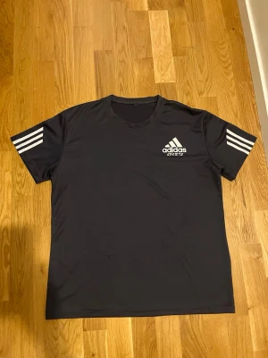 Svart Adidas t-shirt med vita ränder - Snygg svart t-shirt från Adidas med klassiska vita ränder på ärmarna och Adidas-logga på bröstet. Perfekt för sport eller chill. Tillverkad i mjukt och lätt polyester för skön känsla. 🖤