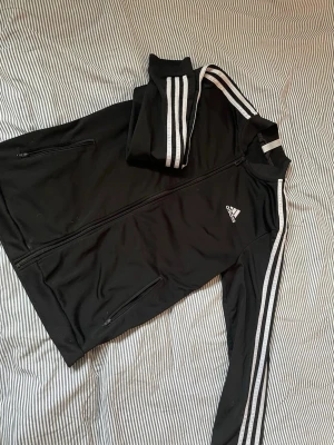 Svart Adidas träningsjacka - Klassisk svart träningsjacka från Adidas med vita ränder längs ärmarna och dragkedja framtill. Jackan har ståkrage, två fickor med dragkedja och Adidas-logga på bröstet. Perfekt för träning eller chill.