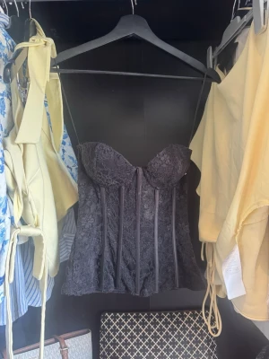 Svart spetskorsett med blomdetaljer - Supersnygg svart korsett med blommig spets och markerade sömmar framtill. Axelbandslös design med vadderade kupor för extra stöd. Perfekt att styla till jeans eller kjol för en trendig look.