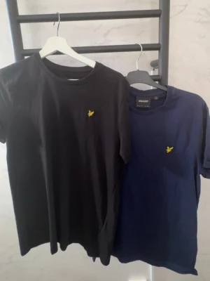 Svart och marinblå t-shirt från lyle and scott - Två stilrena t-shirts i svart och marinblått med gul broderad logga på bröstet. Klassisk rund hals och korta ärmar. Perfekta basplagg för en enkel och snygg look. Tillverkade i mjuk bomull för skön komfort. Båda för 300.
