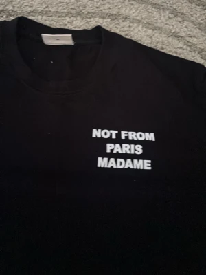 Svart t-shirt med texttryck - Snygg svart t-shirt med vit text 'NOT FROM PARIS MADAME' tryckt både fram och bak. Klassisk rund hals och korta ärmar. Perfekt för dig som gillar statement-plagg och enkel stil.