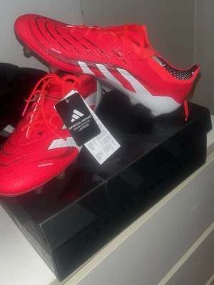 Adidas Predator röda fotbollsskor - Snygga Adidas Predator fotbollsskor i rött med vita och svarta detaljer. Skorna har en modern design med tydliga ränder på sidan, snörning och dobbar för konstgräs. Tillverkade i syntetmaterial för bra grepp och kontroll på planen.