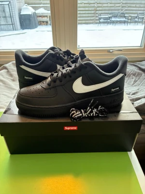 Nike Air Force 1 x Supreme svart/vit - Säljer ett par Nike Air Force 1 Low SP i samarbete med Supreme. Skorna är svarta med vit swoosh och Supreme-logga på sidan. Köpta från supremes webaffär, aldrig använda så e 10/10 skick. 