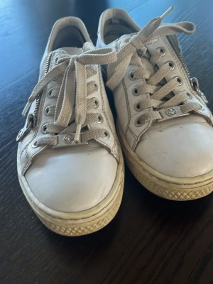 Vita sneakers med dragkedja - Säljer ett par beige sneakers med snörning och dragkedja på sidan. Skorna har en låg profil och en diskret logga på sidan. Ovandelen är i slätt material och sulan är i gummi med rutmönster. Perfekta för en avslappnad stil.