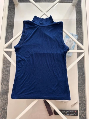 Blå ribbad polotopp ärmlös - Snygg mörkblå ärmlös polotopp i ribbat material. Toppen har en hög krage och är tillverkad i mjuk och stretchig polyester, perfekt för att styla med jeans eller kjol. Enkel men trendig design som passar till många olika looks.