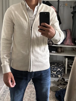 Beige ziptröja med hög krage - Snygg beige ziptröja med hög krage och ribbade muddar. Tröjan har dragkedja framtill och är tillverkad i ett mjukt stickat material. Perfekt att slänga på sig över en skjorta eller t-shirt för en clean look.
