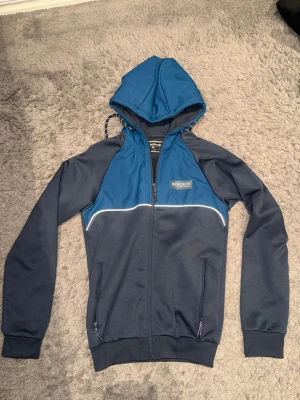 Blå hoodie från McKenzie med dragkedja - Snygg hoodie från McKenzie i två nyanser av blått med dragkedja framtill och huva med dragsko. Har vita detaljer över bröstet och två fickor med dragkedja. Perfekt för en sportig och avslappnad stil.