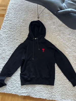 Svart ami paris hoodie - Säljer en svart ami paris hoodie med huva och dragsnören. På bröstet finns ett rött hjärta med bokstaven 'A' broderat. Hoodien har en stor magficka och ribbade muddar vid ärmar och nederkant. Perfekt för en chill och stilren look.