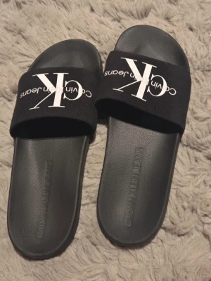 Svarta slides från Calvin Klein Jeans - Svarta slides från Calvin Klein Jeans med bred rem i textil och stor vit CK-logga framtill. Sulan är i svart syntetmaterial och har Calvin Klein Jeans präglat på fotbädden. Perfekta för chill dagar eller till stranden.