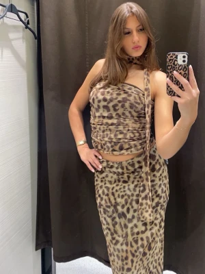 Leopardmönstrad omlottopp med scarf och kjol - Säljer en leopardmönstrad omlottopp och kjol i beige och bruna toner. Toppen har en asymmetrisk design med smala axelband och en matchande scarf som kan knytas runt halsen. Materialet är mjukt och stretchigt, perfekt för en trendig och edgy look.