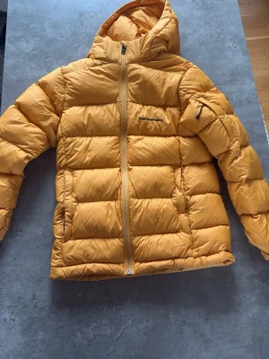 Gul dunjacka, peak performance- ge bud🙌 - ⚠️Kom med bud⚠️ Snygg gul dunjacka med huva och dragkedja framtill. Jackan har quiltad design, två sidofickor och en diskret logga på bröstet samt ärmen. Perfekt för kalla vinterdagar och riktigt varm tack vare dunfyllningen. Gjord för personer i storlek 160cm.