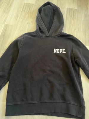 Svart hoodie med tryck 'NOPE.' - Svart hoodie med huva och trycket 'NOPE.' i vitt på bröstet. Klassisk modell med lång ärm och mjukt material. Perfekt för en avslappnad och cool stil.