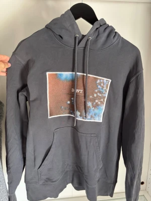 Mörkgrå hoodie med tryck - Säljer en mörkgrå hoodie med huva och dragsnören. På bröstet finns ett rektangulärt tryck i brunt, blått och vitt med texten 'DNT'. Hoodien har en stor magficka och är gjord i mjukt bomullsliknande material. Perfekt för en chill och avslappnad stil.