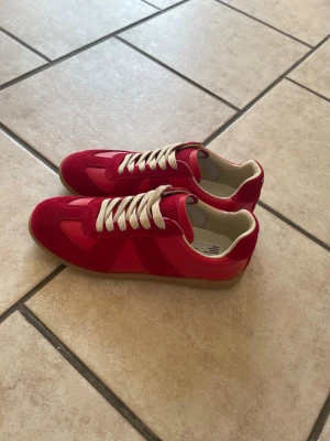 Röda sneakers med beige sula - Säljer ett par snygga röda sneakers med beige sula och vita snören. Skorna har en mix av mocka och slätt material på ovandelen, samt en klassisk låg profil. Perfekta för dig som vill sticka ut med färg på fötterna. Kan gå ner i pris vid snabb affär!