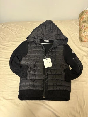 Svart dunväst med huva från Moncler - Snygg svart dunväst från Moncler med stickade ärmar och huva. Västen har dragkedja framtill, två fickor med knapp och Moncler-logga på ärmen. Perfekt för lager-på-lager och streetstyle. Materialet är en mix av dun och stickat tyg.