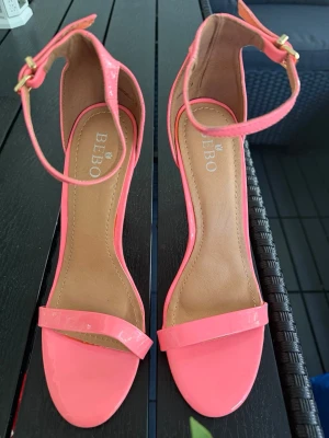 Neonrosa sandaler med ankelrem från BEBO - Säljer ett par snygga neonrosa sandaler från BEBO med öppen tå och smal rem över tårna. Sandalerna har en justerbar ankelrem med guldfärgat spänne och en klack som ger extra höjd. Materialet är glansigt och ger en trendig look.