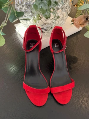 Röda pumps med smal klack - Snygga röda pumps med öppen tå och smal klack. Skorna har en enkel rem över tårna och justerbar ankelrem med spänne. Materialet ser ut att vara mockaimitation och innersulan är svart. Märket är XIT.