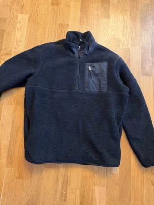 Mörkblå fleece tröja med halv dragkedja - Mysig mörkblå fleece tröja med hög krage och halv dragkedja framtill. Bröstficka med dragkedja och detaljer i matchande tyg. Perfekt för kyliga dagar och enkel att slänga på sig över en t-shirt.