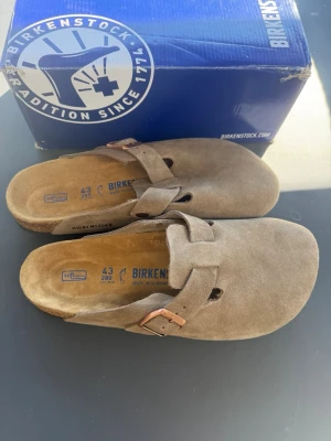 Birkenstock Boston taupe mockatofflor - Säljer ett par Birkenstock Boston tofflor i taupefärgad mocka med justerbart spänne i metall. Klassisk slip-in modell med mjuk fotbädd och rundad tå. Perfekta för chill dagar hemma eller ute på stan.