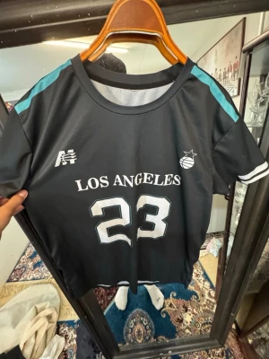 Svart sporttröja Los Angeles 23 - Snygg svart sporttröja med texten 'LOS ANGELES' och siffran 23 på framsidan. Tröjan har vita och turkosa detaljer på axlar och ärmslut samt rund halsringning. Perfekt för basket eller träning.