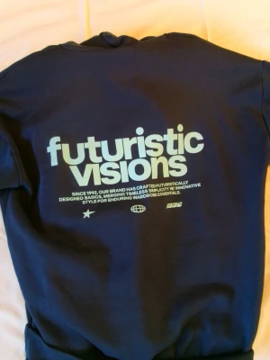 Mörkblå hoodie - Mörkblå hoodie med stor vit text 'futuristic visions' på ryggen. Hoodien har huva och ribbade muddar. Trycket ger en cool och futuristisk känsla.