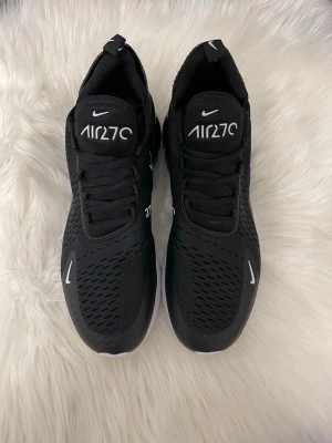 Svarta Nike Air 270 sneakers - Säljer ett par svarta Nike Air 270 sneakers med vit logga och text på plösen. Skorna har en sportig design med meshmaterial för bra ventilation och en tjock, vit sula som ger skön dämpning. Perfekta för dig som gillar streetwear och snygga detaljer.