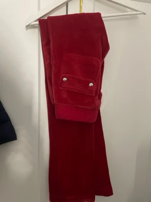 Juicy Couture röda velourbyxor - Snygga röda velourbyxor från Juicy Couture. Byxorna har en ficka med lock och silverfärgade knappar samt dragsko i midjan med metalländar där det står 'Juicy Couture'. Perfekta för en avslappnad men trendig look!