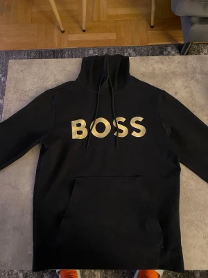 Svart hoodie med guldtext BOSS - Snygg svart hoodie med stor guldtext 'BOSS' på bröstet. Klassisk modell med huva och dragsnören samt en stor magficka framtill. Perfekt för dig som vill sticka ut med en enkel men bold stil.