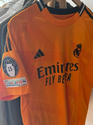Real Madrid matchtröja Vini Jr #7 orange - Säljer en orange Real Madrid matchtröja från Adidas med Vini Jr och nummer 7 på ryggen. Tröjan har svarta detaljer, klubbmärke på bröstet och Champions League-märke på ärmen. Tillverkad i lätt och ventilerande material, perfekt för fotbollsträning eller match.