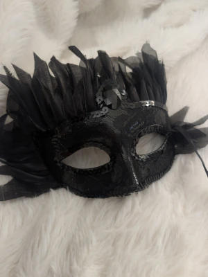 Svart maskeradmask med fjädrar - Snygg svart maskeradmask dekorerad med svarta fjädrar, paljetter och en stor svart sten framtill. Masken har spetsdetaljer och knyts med band. Perfekt till maskerad, cosplay eller temafest.