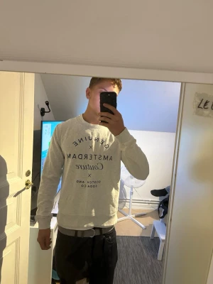 Scotch & Soda Amsterdam Couture Sweatshirt - Säljer en ljusgrå sweatshirt med blått texttryck på bröstet där det står 'Genuine Amsterdam Couture'. Perfekt för en avslappnad och stilren look.