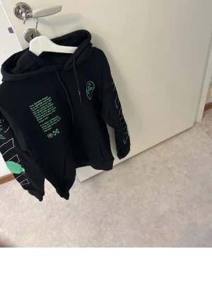 Svart hoodie med grönt tryck - Svart hoodie med huva och gröna grafiska tryck samt textdetaljer på både framsidan och ärmen. Stora Off-White-inspirerade motiv på ryggen och bröstet. Känguruficka framtill och snören i huvan.