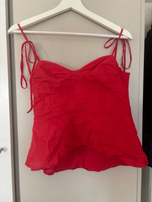 Röd topp med knytband - Snygg röd topp med tunna axelband som knyts på axlarna. Modellen har en lätt rynkad effekt över bysten och är något utsvängd nedtill. Perfekt för sommaren och enkel att styla till jeans eller kjol.