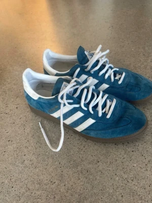 Adidas blå Gazelle sneakers - Säljer ett par blå Adidas Gazelle sneakers med klassiska vita ränder och vit häl. Skorna har ovandel i mocka, vita skosnören och gummisula. Perfekta för dig som gillar retrostil och streetwear.