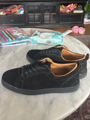 Svarta sneakers i mocka - Säljer ett par stilrena svarta christian louboutin i mocka med svarta skosnören och diskret broderad text på hälen. Skorna har en låg profil och innersida i beige läderimitation. Perfekta för dig som gillar en clean look.