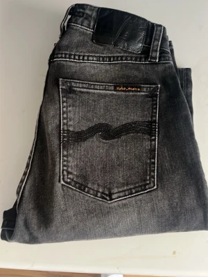Nudie jeans  - Säljer ett par mörk gråa Nudie Jeans. Jeansen har hög midja och raka ben. Midja 30cm, längd 102cm. JAG SKICKAR EJ FLER BILDER NÄR JAG BÄR JEANSEN! för bättre inblick i passformen rekommenderar jag att googla på modellnamnet. För storleksguide kolla måtten noggrant då jeansen alltid kan vara uppsydda heller krympta och jag ej tar emot returer :) (n7)