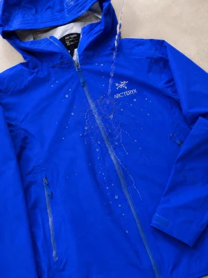 Blå regnjacka från Arc'teryx - Snygg blå regnjacka från Arc'teryx med huva och hel dragkedja framtill. Jackan har två sidofickor med dragkedja och justerbara ärmslut med kardborre. Tillverkad i ett lätt och vattentätt material som håller dig torr även i ösregn.