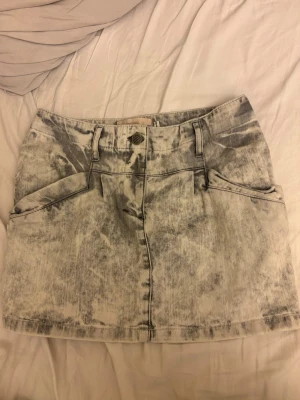 Ljusgrå jeanskjol - Cool kort jeanskjol i acid wash med grå och vita toner. Kjolen har fickor fram och bak, bälteshällor och knappstängning. Perfekt för dig som gillar en trendig och avslappnad stil.