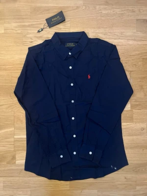 Mörkblå skjorta från Polo Ralph Lauren - Klassisk mörkblå skjorta från Polo Ralph Lauren med vit knappstängning och den ikoniska röda logotypen broderad på bröstet. Skjortan har lång ärm och är tillverkad i mjuk bomull. Perfekt för en stilren look. Inte använts så många gånger, inga defekter eller så. Storlek M, men sitter som S. Säljer pågrund av att den är för liten. Hör av dig vid frågor.