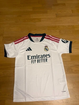 Real Madrid hemma 26/27 S - Snygg vit Real Madrid matchtröja från Adidas med klubbmärke och sponsortryck på bröstet. Tröjan har mörkblå detaljer vid krage och ärmslut samt rosa ränder på axlarna. Tillverkad i lätt och ventilerande material, perfekt för fotbollsträning eller match.