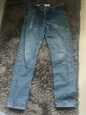 Blå Jack&Jones Jeans - Säljer ett par blå jeans med rak passform i storlek 28/32 och hög midja. Jeansen har fem fickor, knappgylf och subtila slitningar framtill. Tillverkade i kraftigt denimtyg som ger en tidlös look. Perfekta till en avslappnad stil.