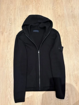 Stone Island cardigan - Stone Island cardigan. Tröjan har en ribbstickad struktur och en patch på vänster ärm. Perfekt för en avslappnad och stilren look.
